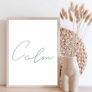Op de afbeelding: Een ingelijst print met het woord "Calm" geschreven in een cursief schrift in lichtblauw op een witte achtergrond. De print staat op een houten oppervlak naast een vaas met gedroogd pampasgras.