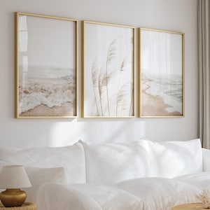 Op de afbeelding: Een woonkamer met een witte bank, twee witte ronde stoelen en twee ronde houten salontafels. Drie ingelijste prints met gouden randen met strandscènes en pampasgras hangen aan de muur boven de bank. Een rond jute kleed ligt op de vloer.