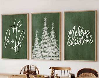 Pode incluir: Três impressões de tela verde emolduradas com letras brancas e uma silhueta branca de árvores de Natal. As impressões estão penduradas em uma parede branca acima de uma mesa de jantar de madeira com cadeiras de vime.