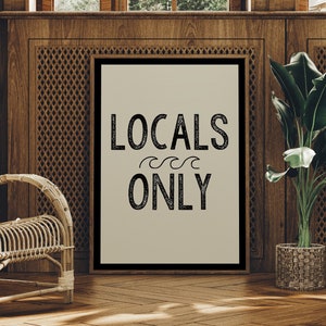 May include: A framed print with the text "LOCALS ONLY" in black lettering on a beige background. The text is stylized with a wavy line underneath the word "LOCALS". The print is hanging on a wooden wall with a wicker chair and a potted plant in the foreground.