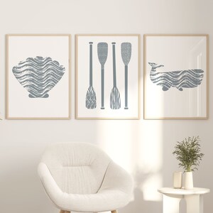 Puede incluir: Tres impresiones enmarcadas con un tema n&aacute;utico. Las impresiones presentan una concha, tres remos y una ballena. Las impresiones est&aacute;n colgadas en una pared blanca sobre un sill&oacute;n blanco y una mesa auxiliar blanca.