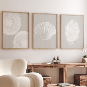 K&ouml;nnte beinhalten: Drei gerahmte Drucke mit wei&szlig;en Illustrationen einer Muschel, einer Koralle und einem kreisf&ouml;rmigen Muster auf hellgrauem Hintergrund. Die Drucke h&auml;ngen an einer wei&szlig;en Wand &uuml;ber einem Holztisch mit einem wei&szlig;en Sessel und einem Holz-Couchtisch.