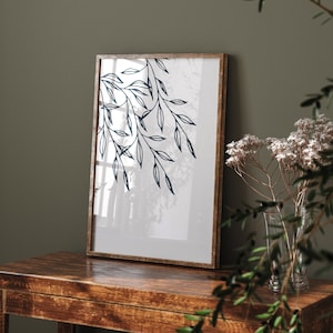 Puede incluir: Una impresión enmarcada de un diseño floral estilizado en blanco y negro sobre un fondo blanco. El marco es de madera marrón y la impresión está sobre una mesa de madera marrón oscura.