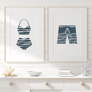 Op de afbeelding: Twee ingelijste prints van een badpak en een zwembroek. Het badpak is een zwart-wit gestreept bikini-topje en -onderdeel. De zwembroek is zwart-wit gestreept met een wit trekkoord. De prints zijn op een witte achtergrond en zijn ingelijst in een wit frame.