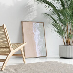 May include: A framed photograph of an aerial view of a beach with waves crashing on the shore. The photo is hanging on a white wall next to a woven rattan chair and a potted palm tree.