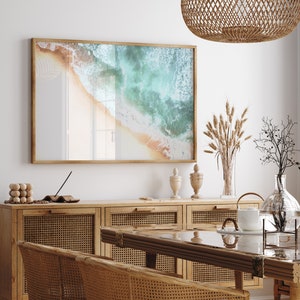 May include: A framed photograph of a beach scene with turquoise water and white sand. The photo is hanging on a white wall above a wooden cabinet with woven rattan doors. There are decorative items on the cabinet, including a vase with dried wheat stalks and a glass vase with a branch.