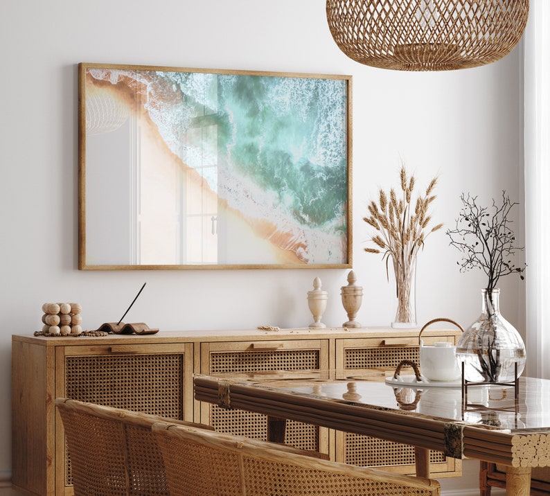 May include: A framed photograph of a beach scene with turquoise water and white sand. The photo is hanging on a white wall above a wooden cabinet with woven rattan doors. There are decorative items on the cabinet, including a vase with dried wheat stalks and a glass vase with a branch.