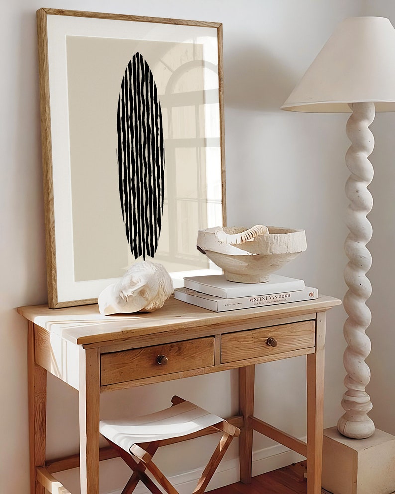 Puede incluir: Una impresi&oacute;n enmarcada con un dise&ntilde;o abstracto en blanco y negro de una tabla de surf sobre un fondo beige. La impresi&oacute;n est&aacute; colgada en una pared sobre una mesa de madera con dos cajones. Un cuenco de cer&aacute;mica blanco y una escultura blanca est&aacute;n sobre la mesa.