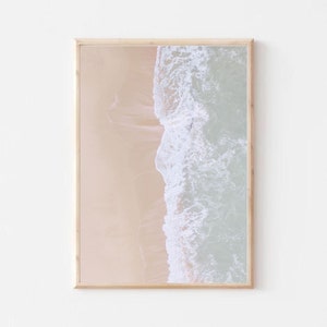 May include: An aerial photograph of a sandy beach with a wave crashing on the shore. The water is a light blue and the sand is a light beige.