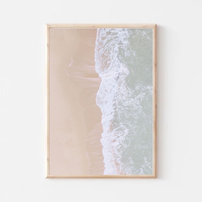 May include: An aerial photograph of a sandy beach with a wave crashing on the shore. The water is a light blue and the sand is a light beige.