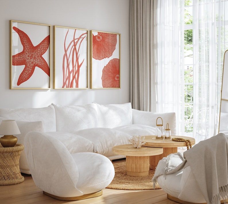 May include: A white sectional sofa with a white throw pillow, a white armchair, and a wooden coffee table with a white coral sculpture on top. The room has a white wall with three framed prints of red coral and starfish. The room has a large window with white curtains.