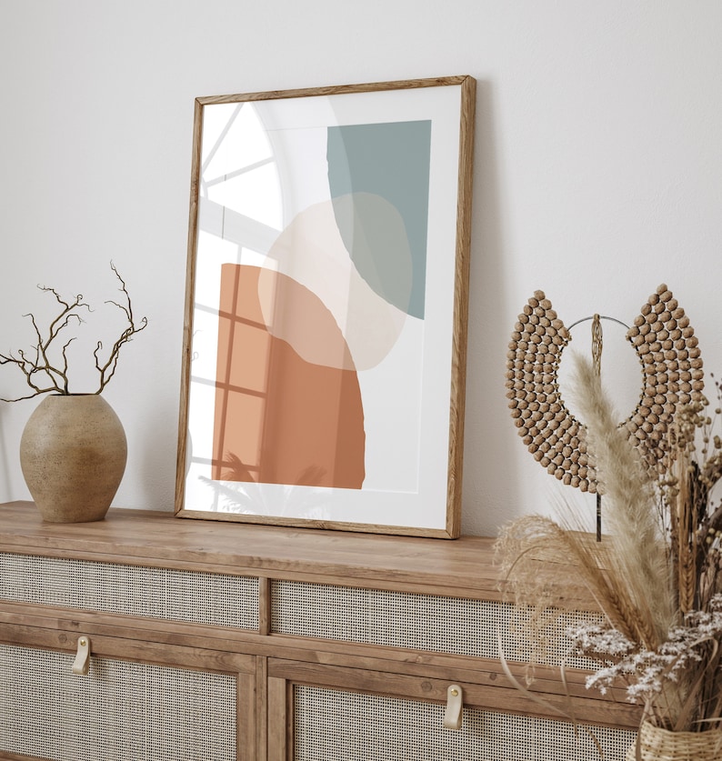 May include: A framed abstract print featuring geometric shapes in shades of orange, brown, and blue. The print is hanging on a white wall above a wooden dresser with woven rattan drawer fronts.