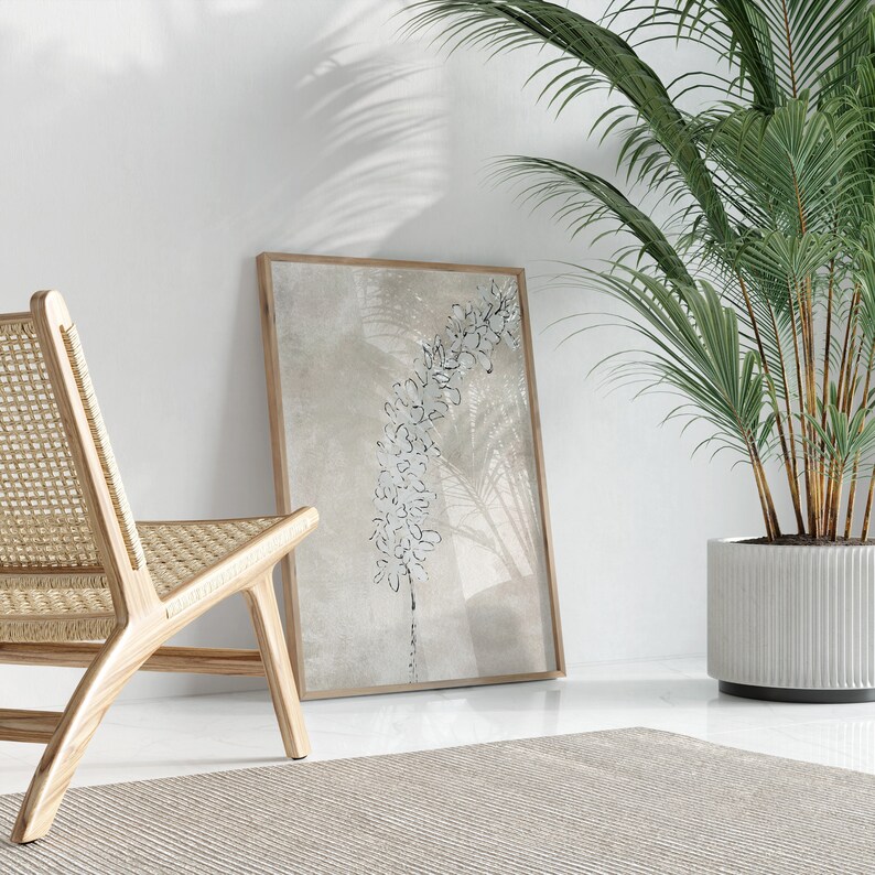 Puede incluir: Una impresi&oacute;n enmarcada de un dise&ntilde;o floral estilizado en blanco y negro sobre un fondo beige. La impresi&oacute;n est&aacute; colgada en una pared blanca junto a una silla de mimbre y una palmera en maceta.