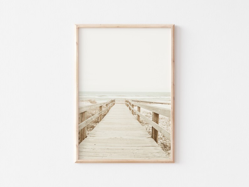 May include: A wooden boardwalk leads to the ocean, with a light brown wooden railing on either side. The ocean is a light blue color, and the sky is a light gray color.
