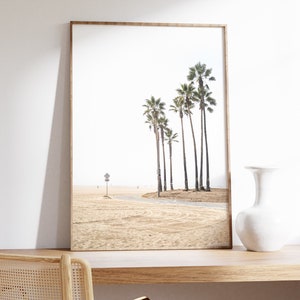 May include: A framed print of a beach scene with palm trees in the distance. The beach is sandy and there is a road leading to the trees. The print is in a light wood frame.