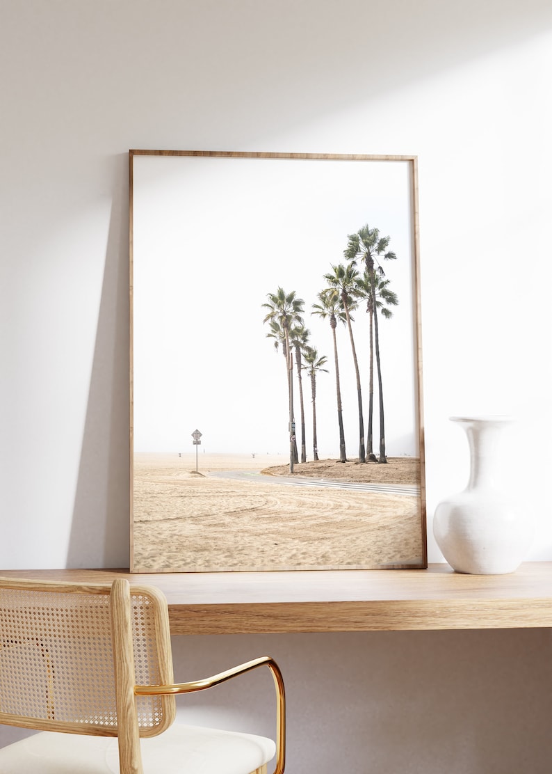 May include: A framed print of a beach scene with palm trees in the distance. The beach is sandy and there is a road leading to the trees. The print is in a light wood frame.