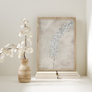Puede incluir: Una impresi&oacute;n enmarcada de una ilustraci&oacute;n bot&aacute;nica en blanco y negro de una flor sobre un fondo beige. La impresi&oacute;n est&aacute; sobre una pila de libros sobre una superficie blanca. Un jarr&oacute;n con una rama frondosa est&aacute; en primer plano.