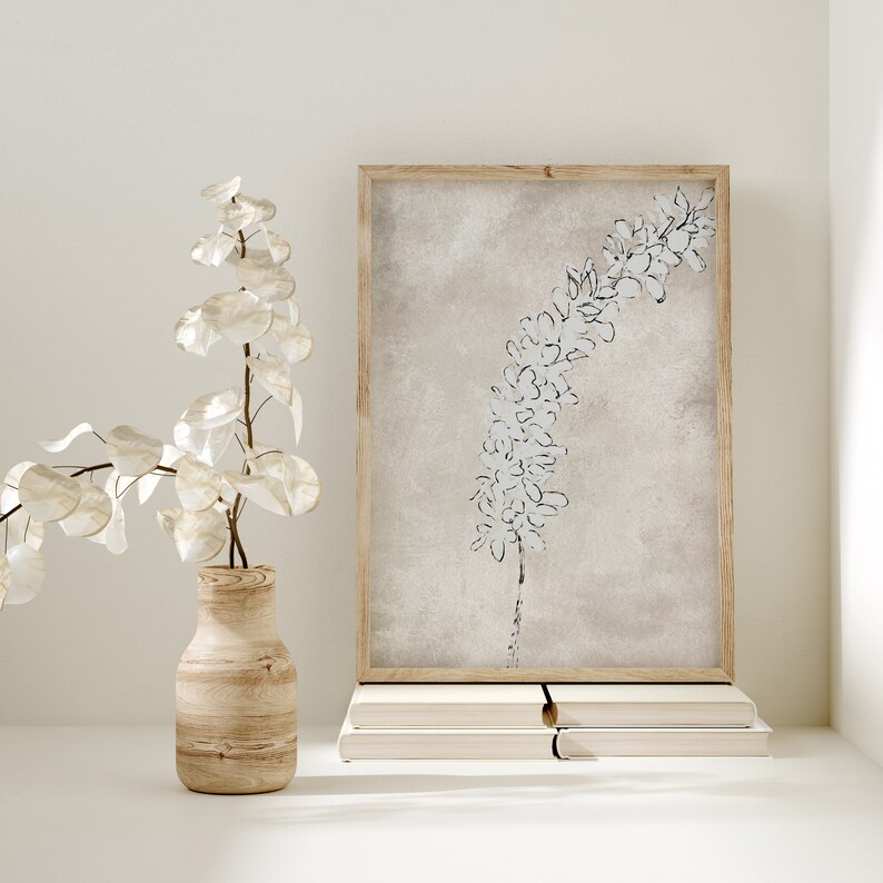 Puede incluir: Una impresi&oacute;n enmarcada de una ilustraci&oacute;n bot&aacute;nica en blanco y negro de una flor sobre un fondo beige. La impresi&oacute;n est&aacute; sobre una pila de libros sobre una superficie blanca. Un jarr&oacute;n con una rama frondosa est&aacute; en primer plano.