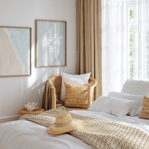 May include: A white bed with a beige knitted throw blanket and a straw hat. The bed is in a bedroom with white walls, a wicker chair with a beige pillow, and two framed prints of palm trees and a beach scene.