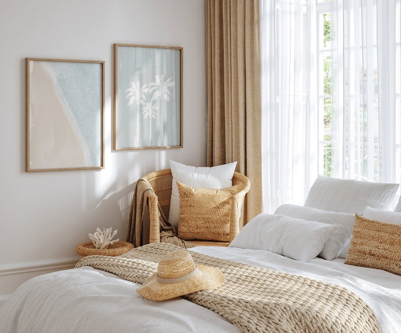 May include: A white bed with a beige knitted throw blanket and a straw hat. The bed is in a bedroom with white walls, a wicker chair with a beige pillow, and two framed prints of palm trees and a beach scene.