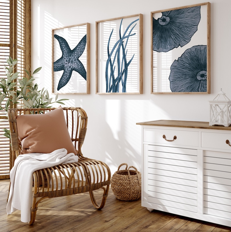 Op de afbeelding: Een wit kastje met twee lades en twee deuren, een rieten stoel met een bruin kussen, een geweven mand en drie ingelijste prints van blauwe zeesterren, zeewier en koraal op een witte muur.