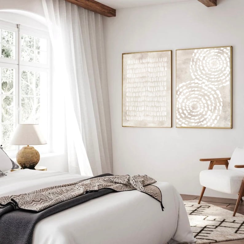 Puede incluir: Dos impresiones de arte abstracto en marcos dorados con dise&ntilde;os geom&eacute;tricos blancos sobre un fondo beige. Las impresiones est&aacute;n colgadas en una pared blanca en un dormitorio con una cama blanca y una silla blanca.