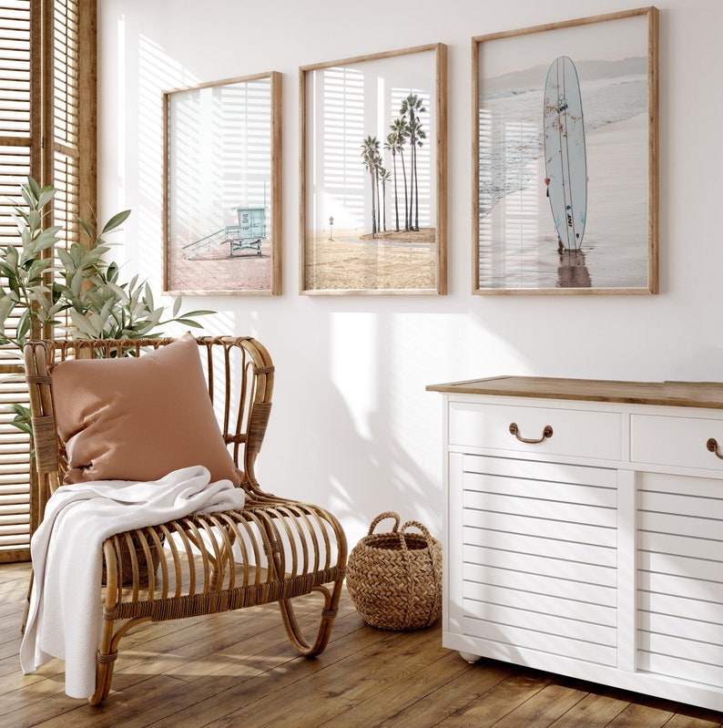 May include: A wicker chair with a brown throw blanket and a tan cushion sits in a room with three framed beach-themed prints and a white cabinet with two drawers.