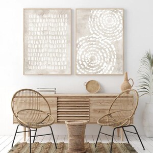 Puede incluir: Dos impresiones de arte abstracto enmarcadas con dise&ntilde;os geom&eacute;tricos blancos sobre un fondo beige. Las impresiones est&aacute;n colgadas en una pared blanca sobre una consola de madera con dos sillas de mimbre y un taburete de mimbre.