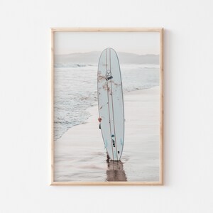 May include: A white surfboard with a blue and black logo is standing on a sandy beach with ocean waves in the background.