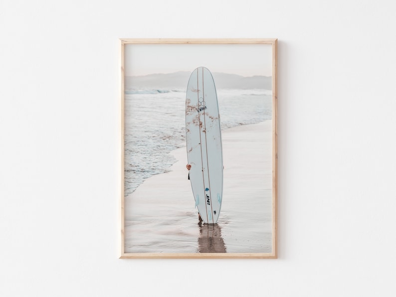May include: A white surfboard with a blue and black logo is standing on a sandy beach with ocean waves in the background.
