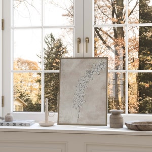 Puede incluir: Un dibujo en blanco y negro de una rama con hojas sobre un fondo beige, enmarcado en un marco dorado. El dibujo est&aacute; colocado sobre un alf&eacute;izar de ventana blanco con una vela, un jarr&oacute;n peque&ntilde;o y un cuenco de madera.