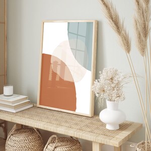 May include: A framed abstract print with a white background and overlapping shapes in shades of orange, brown, and blue. The print is hanging on a wall above a wooden bench with a woven rattan top.