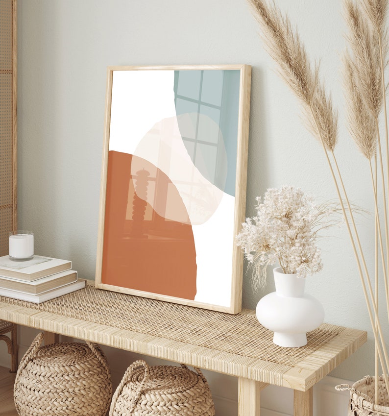 May include: A framed abstract print with a white background and overlapping shapes in shades of orange, brown, and blue. The print is hanging on a wall above a wooden bench with a woven rattan top.