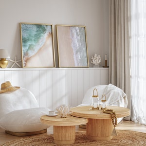 May include: Two framed beach scene prints with a white, gold, and beige color scheme hang above a white wainscoting wall. Two white chairs with a throw blanket on one are positioned in front of a round, natural wood coffee table with a smaller round table nested underneath. The coffee table has a gold candle holder and a beaded garland.