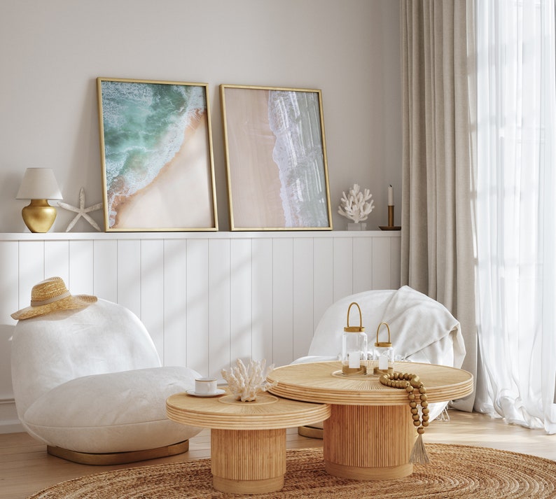 May include: Two framed beach scene prints with a white, gold, and beige color scheme hang above a white wainscoting wall. Two white chairs with a throw blanket on one are positioned in front of a round, natural wood coffee table with a smaller round table nested underneath. The coffee table has a gold candle holder and a beaded garland.