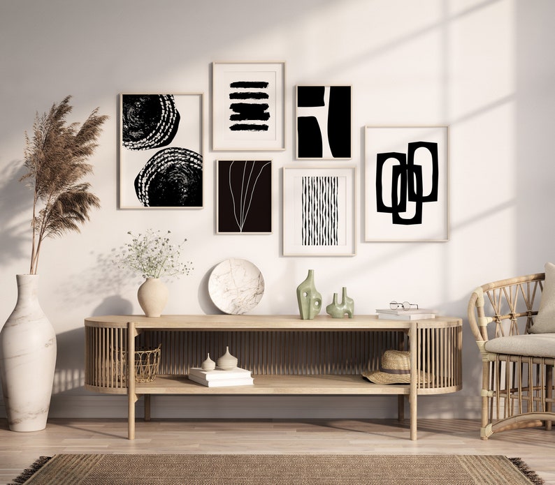 May include: A living room interior with a light wood console table featuring a collection of black and white abstract art prints. The console table has two shelves and is decorated with a vase of white flowers, a ceramic plate, and two small green vases. A wicker chair with a white cushion is in the background.
