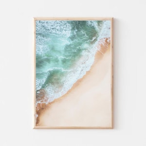 May include: An aerial view of a sandy beach with turquoise ocean waves crashing on the shore. The water is a vibrant blue-green color, and the sand is a light beige.