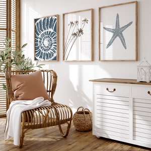 May include: A white wicker chair with a brown cushion and a white throw blanket. The chair is in front of a white wall with three framed prints of a seashell, palm trees, and a starfish. A white cabinet with a wooden top is in the background.