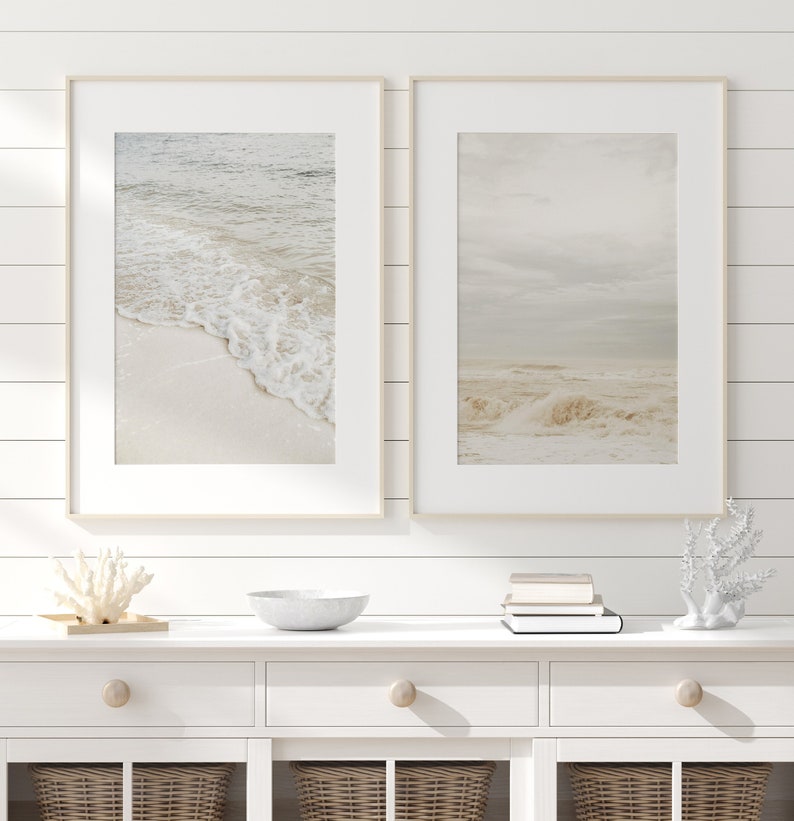May include: Two framed prints of seascapes with white sand and foamy waves. The prints are hung on a white shiplap wall above a white dresser with three drawers.