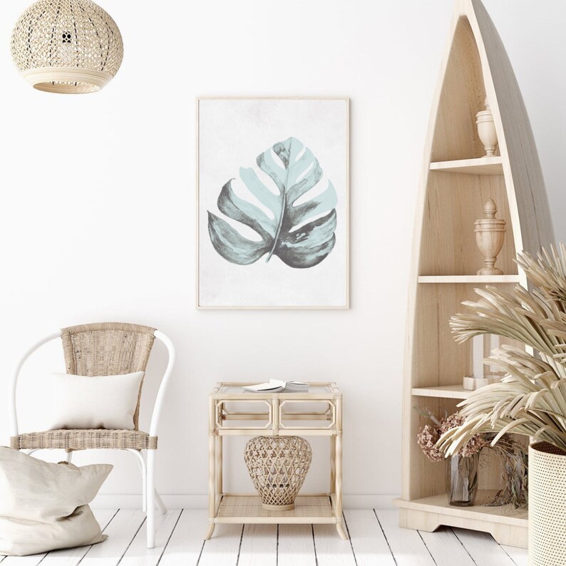 May include: A white wicker chair with a white pillow, a light wood side table with a woven basket, and a framed print of a light blue and gray tropical leaf on a white background.