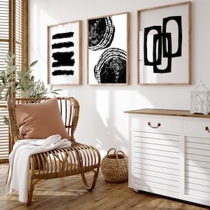 May include: Three black and white abstract art prints in wooden frames hanging on a white wall above a white cabinet with a wooden top. A wicker chair with a brown pillow and a white throw blanket sits in front of the cabinet.