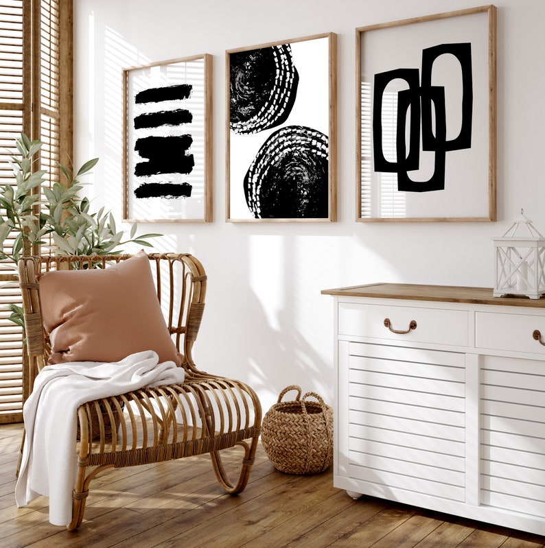 May include: Three black and white abstract art prints in wooden frames hanging on a white wall above a white cabinet with a wooden top. A wicker chair with a brown pillow and a white throw blanket sits in front of the cabinet.