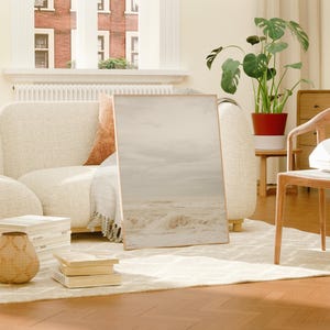 May include: A white couch with a throw blanket and a framed print of a beach scene. A wooden chair with a stack of books sits in the foreground.