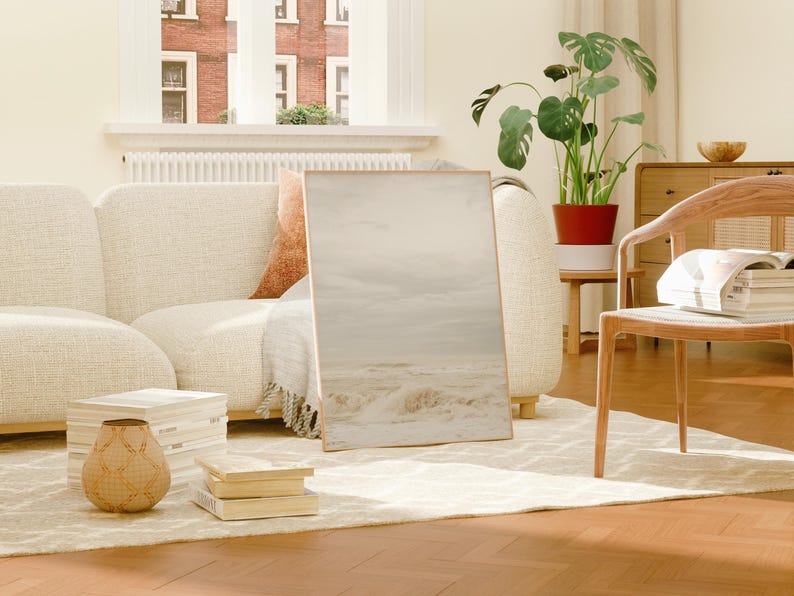 May include: A white couch with a throw blanket and a framed print of a beach scene. A wooden chair with a stack of books sits in the foreground.