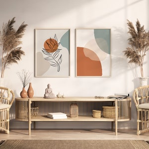 May include: A wooden console table with two shelves, featuring a minimalist design with a natural wood finish. The table is decorated with two vases, a small Buddha statue, and two framed abstract art prints with geometric shapes and a leaf design in shades of brown, orange, and blue. The table is flanked by two wicker chairs with white cushions.