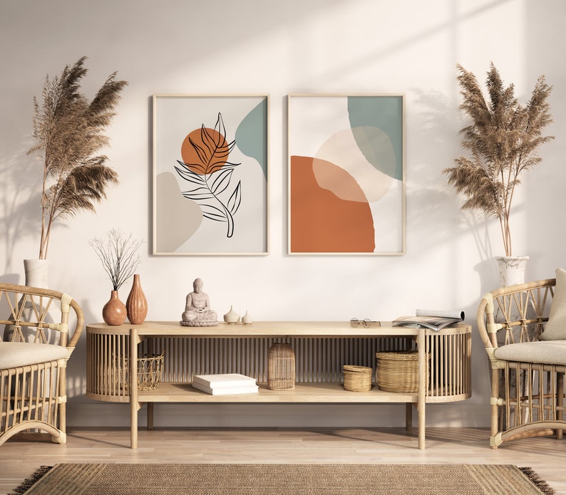 May include: A wooden console table with two shelves, featuring a minimalist design with a natural wood finish. The table is decorated with two vases, a small Buddha statue, and two framed abstract art prints with geometric shapes and a leaf design in shades of brown, orange, and blue. The table is flanked by two wicker chairs with white cushions.