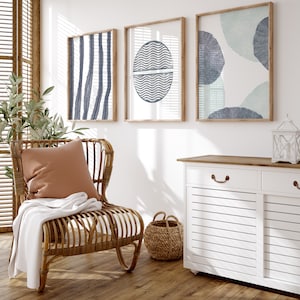 May include: A wicker chair with a brown cushion and a white throw blanket sits in a room with three framed abstract prints featuring blue and green geometric shapes. A white cabinet with a wood top and two drawers is in the background.