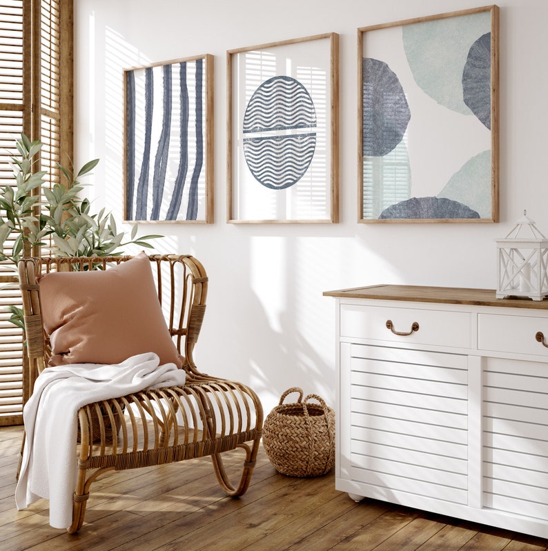 May include: A wicker chair with a brown cushion and a white throw blanket sits in a room with three framed abstract prints featuring blue and green geometric shapes. A white cabinet with a wood top and two drawers is in the background.