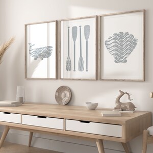 Puede incluir: Tres impresiones enmarcadas con un tema n&aacute;utico. Las impresiones presentan una ballena, remos y una concha, todo en un color azul claro. Las impresiones est&aacute;n colgadas en una pared blanca sobre una mesa consola de madera.
