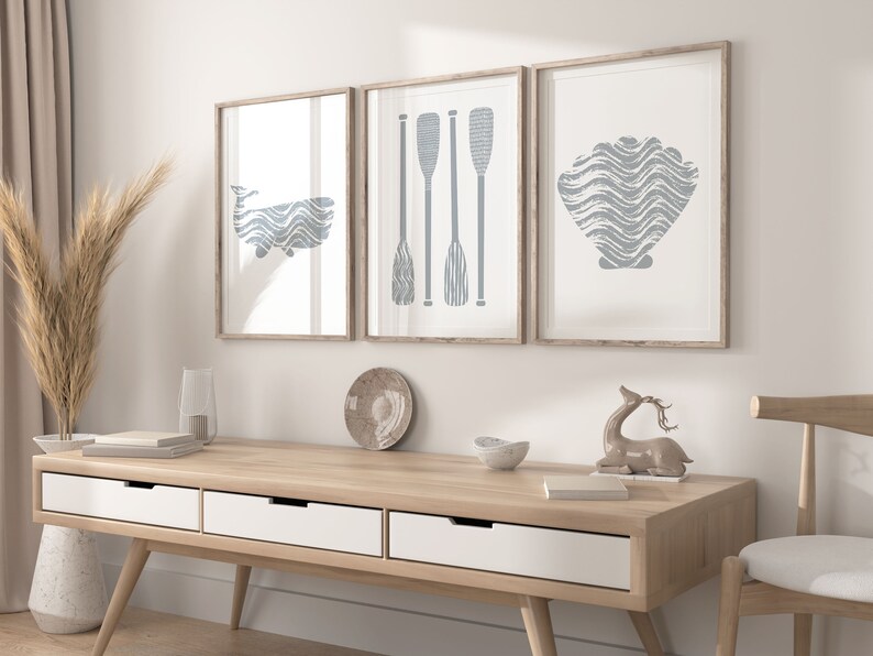 Puede incluir: Tres impresiones enmarcadas con un tema n&aacute;utico. Las impresiones presentan una ballena, remos y una concha, todo en un color azul claro. Las impresiones est&aacute;n colgadas en una pared blanca sobre una mesa consola de madera.